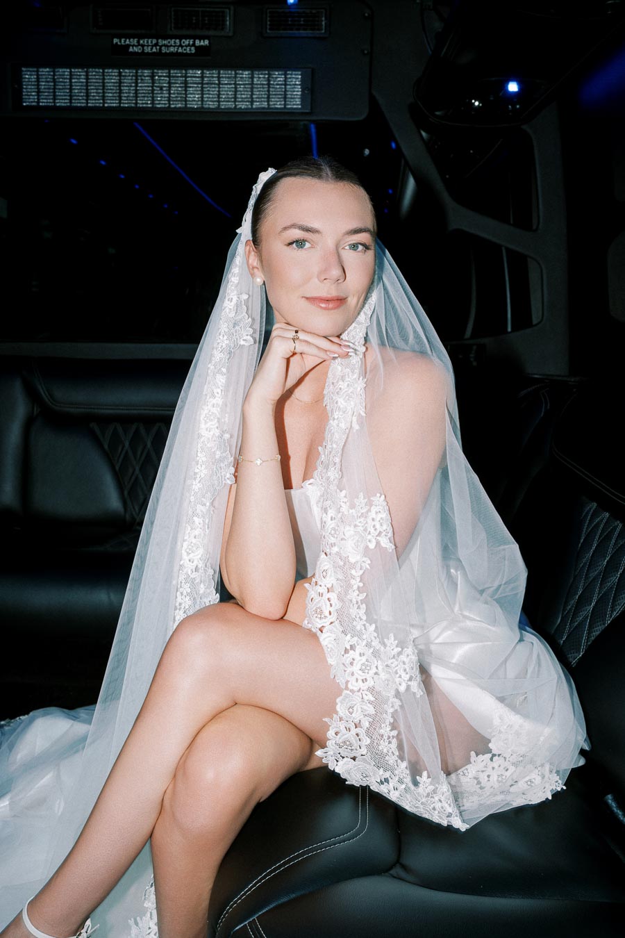 A bride elegantly posing in a luxurious car, wearing a detailed white lace wedding veil.