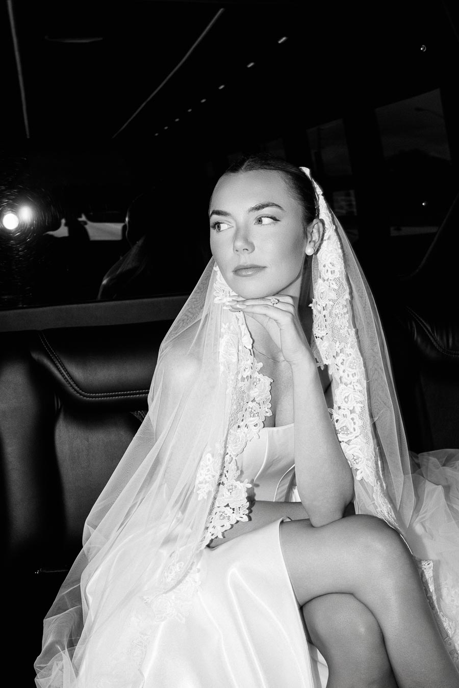 Black and white photo of a bride in an elegant lace veil and satin dress, seated and gazing thoughtfully, perfect for wedding inspiration and bridal fashion.