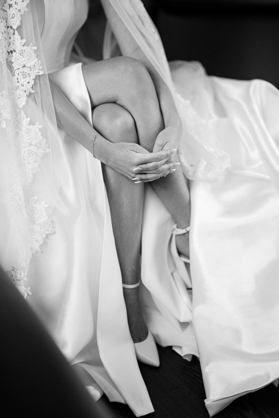 Black and white photo of a bride in an elegant gown, sitting with hands clasped on her knees, showcasing the detailed lace and satin fabric.