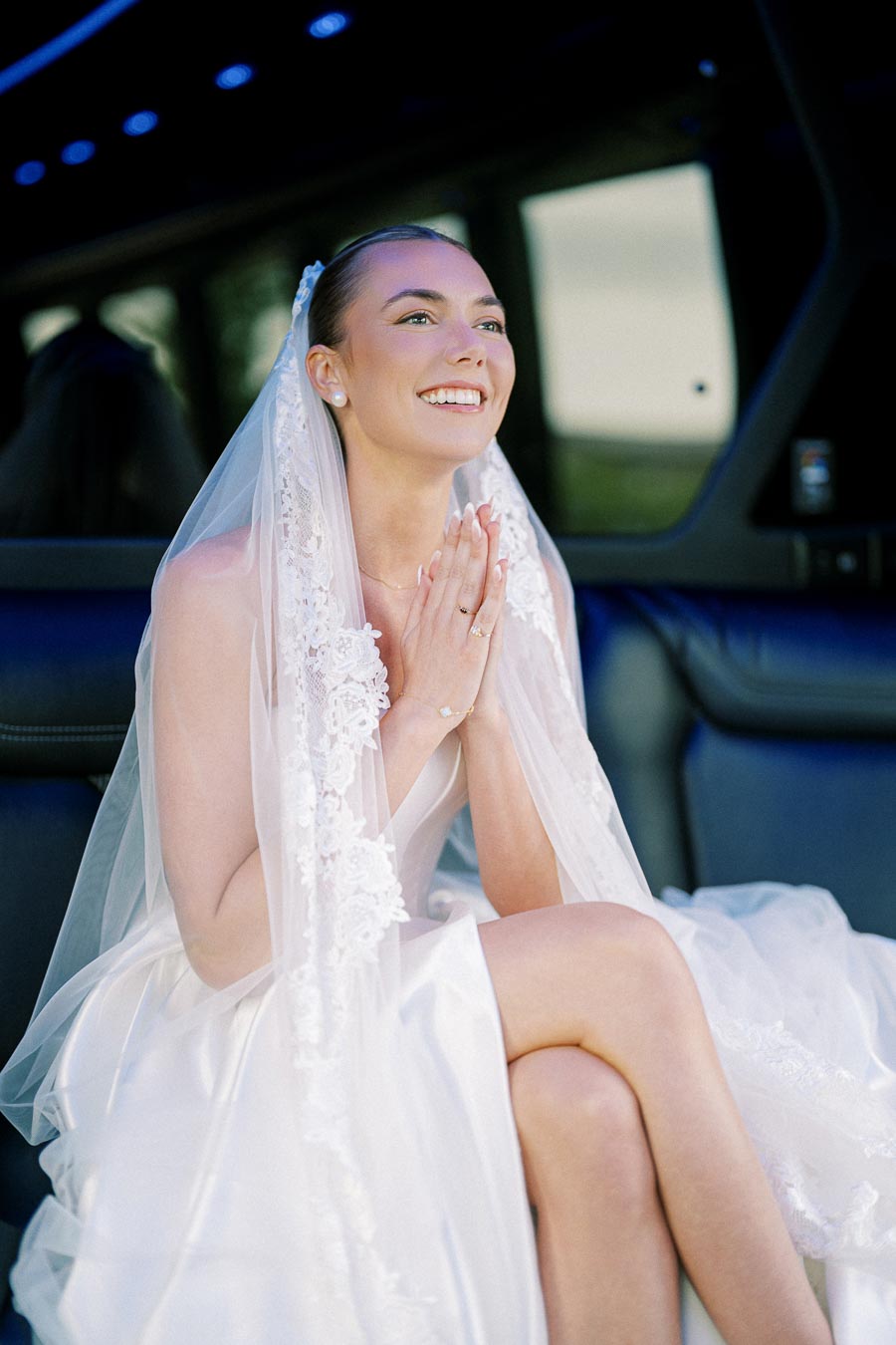 A joyful bride in a white wedding dress and lace veil, sitting in a limousine, looking upwards with hands clasped.