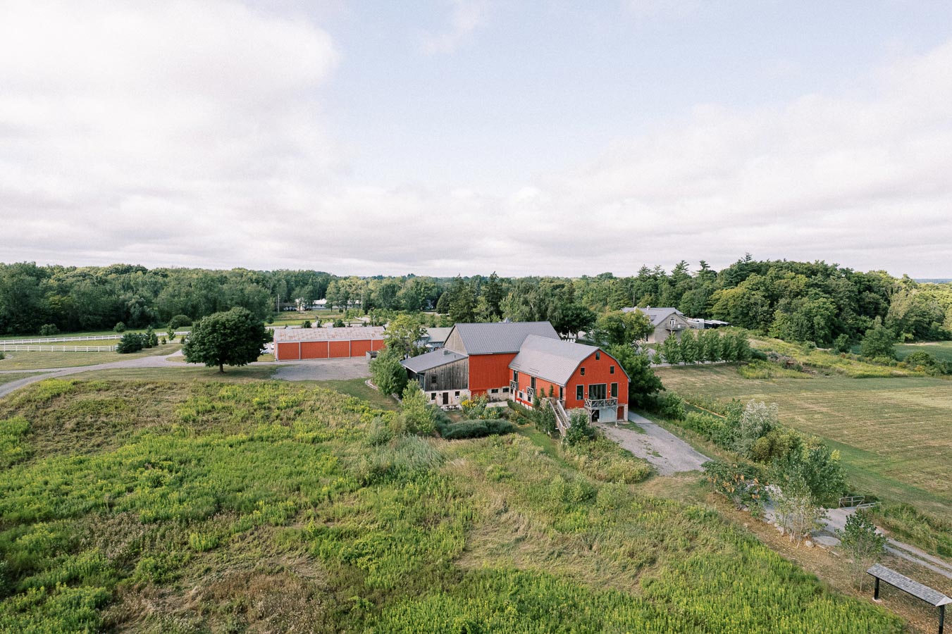 Aerial view of a rural landscape featuring a traditional red barn surrounded by lush greenery and fields, under a partly cloudy sky.