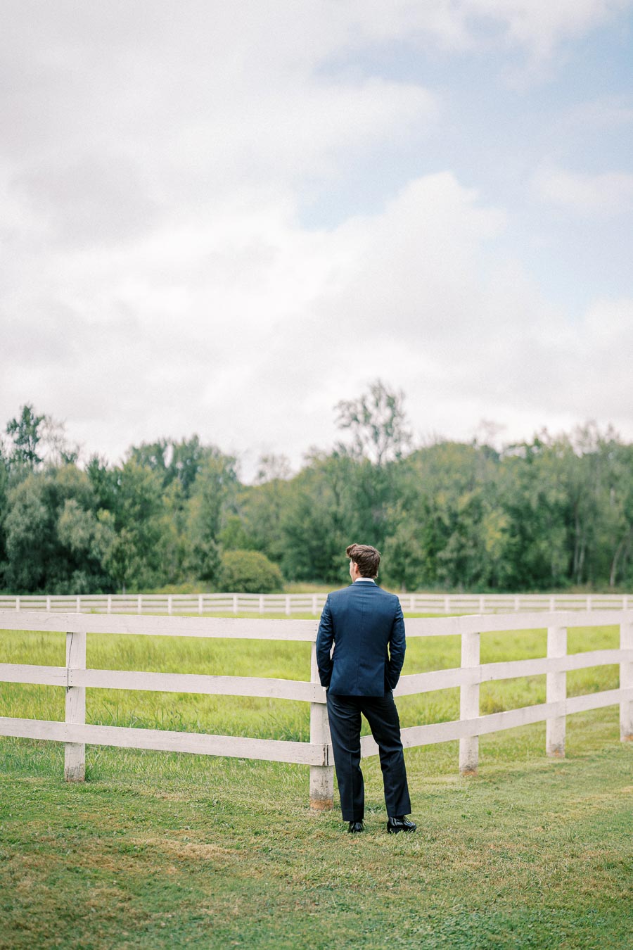 A man in a blue suit stands by a white wooden fence, overlooking a lush green pasture and forest under a cloudy sky.