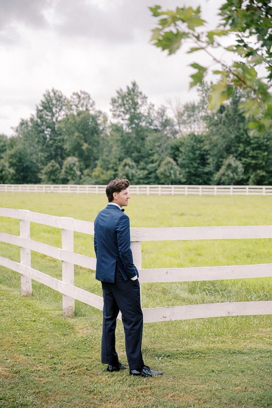 Man in a blue suit standing by a white wooden fence in a grassy field with trees in the background, under a cloudy sky.