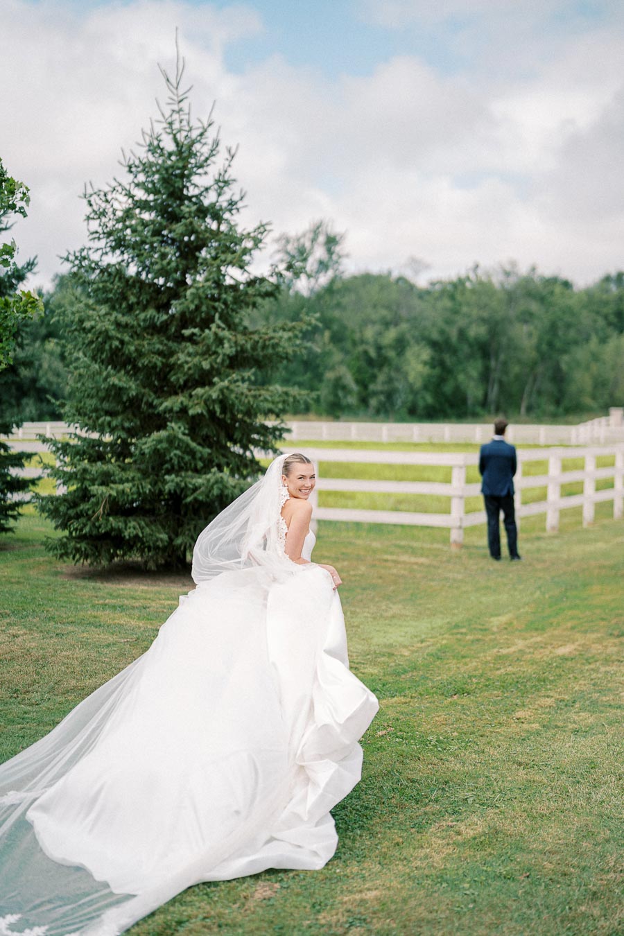 A bride in a flowing white wedding gown and veil smiles while walking outdoors on a grassy field, with a background of green trees and a wooden fence, as a groom stands at a distance.
