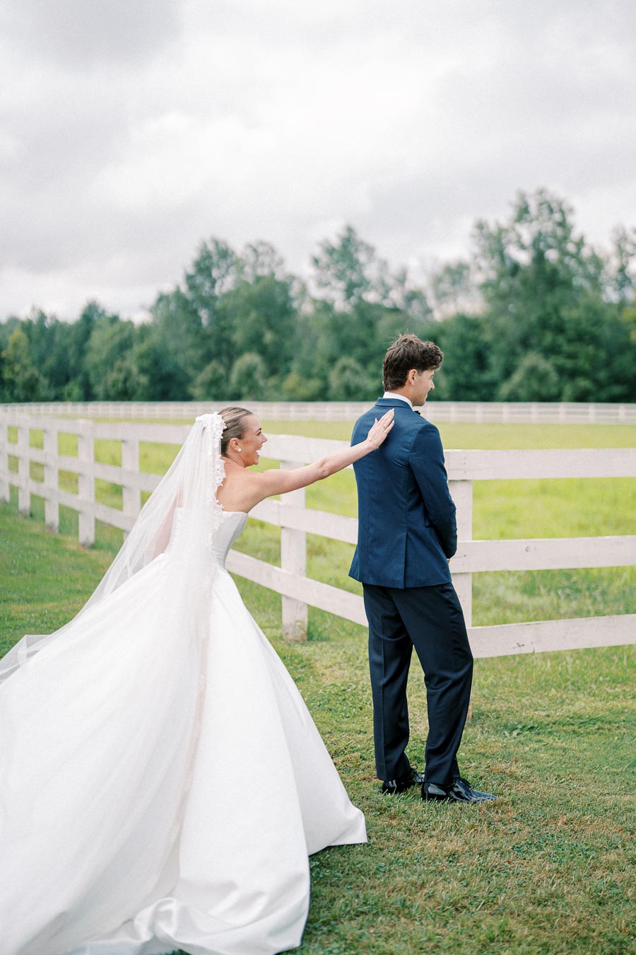 Bride in elegant white gown playfully taps groom on the shoulder for a romantic first-look moment in a picturesque outdoor setting with a white fence and greenery.