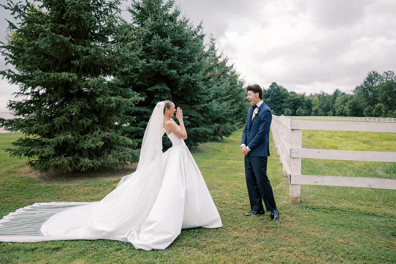 Bride and groom sharing a joyful first look moment outdoors near a white fence and evergreen trees before their wedding ceremony, with the bride in a white gown and veil, and the groom in a blue suit.