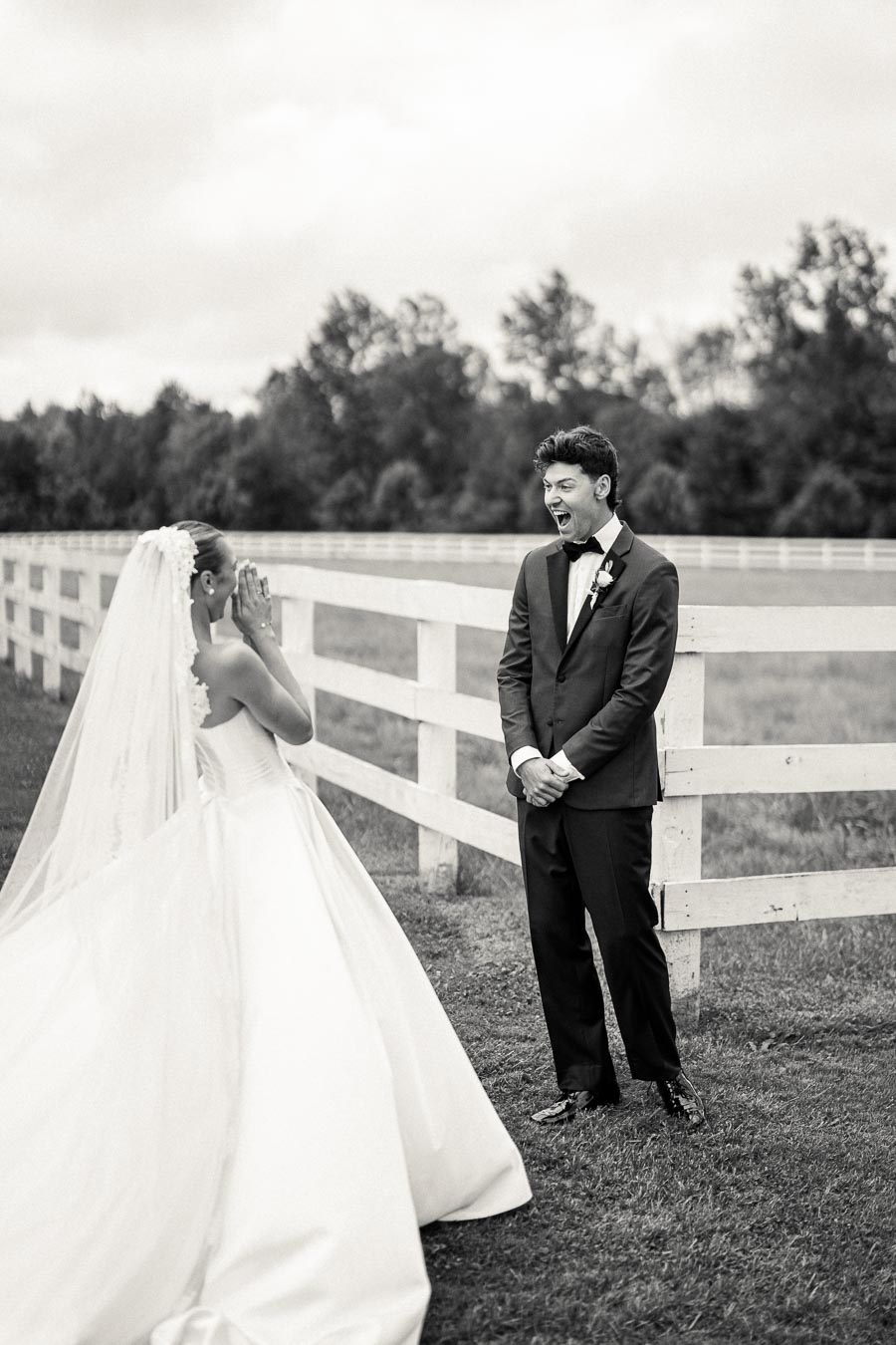 A bride in a white wedding gown surprises the groom in a tuxedo during a first look moment, standing in front of a white fence in an outdoor setting.