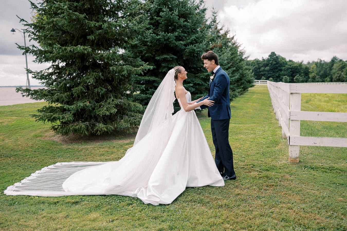 Elegant outdoor wedding scene with a bride in a flowing white gown and veil holding hands with a groom in a navy suit, standing beside lush green trees and a white wooden fence under a cloudy sky.