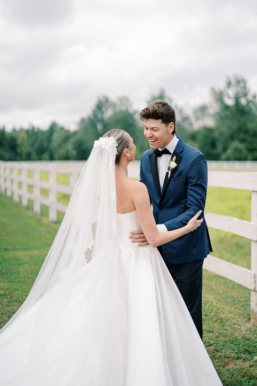 Bride and groom smiling and embracing in front of a white fence and lush greenery, bride wearing elegant white dress and veil, groom in a stylish blue suit.