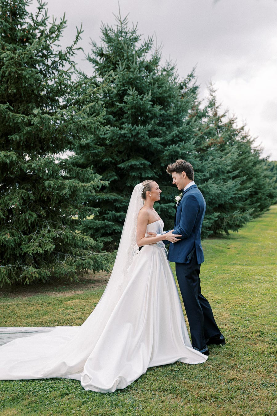 A bride in a white gown with a long train and a groom in a navy suit stand lovingly in front of lush evergreen trees on a wedding day, set against a cloudy sky.