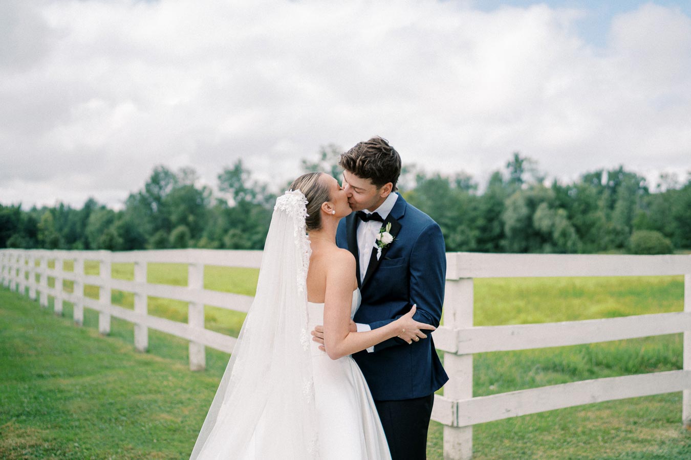 A bride and groom kissing in front of a white fence in a lush green field, with the bride wearing a white wedding dress and veil and the groom in a navy blue suit on a cloudy day.
