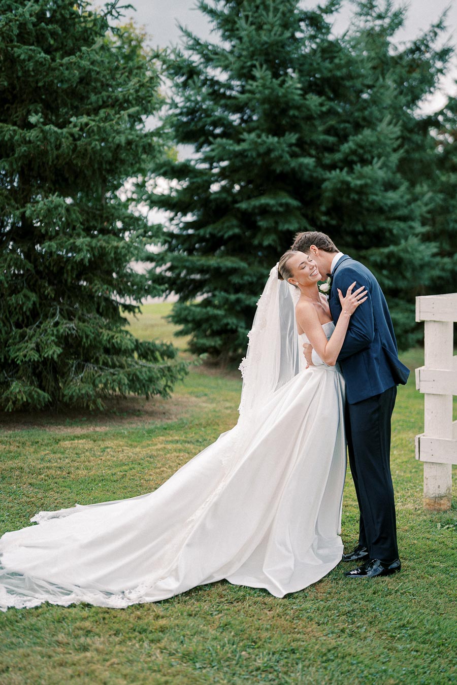 Outdoor wedding portrait of a bride in a flowing white gown and groom in a dark suit, embracing under evergreen trees on a lush green lawn.