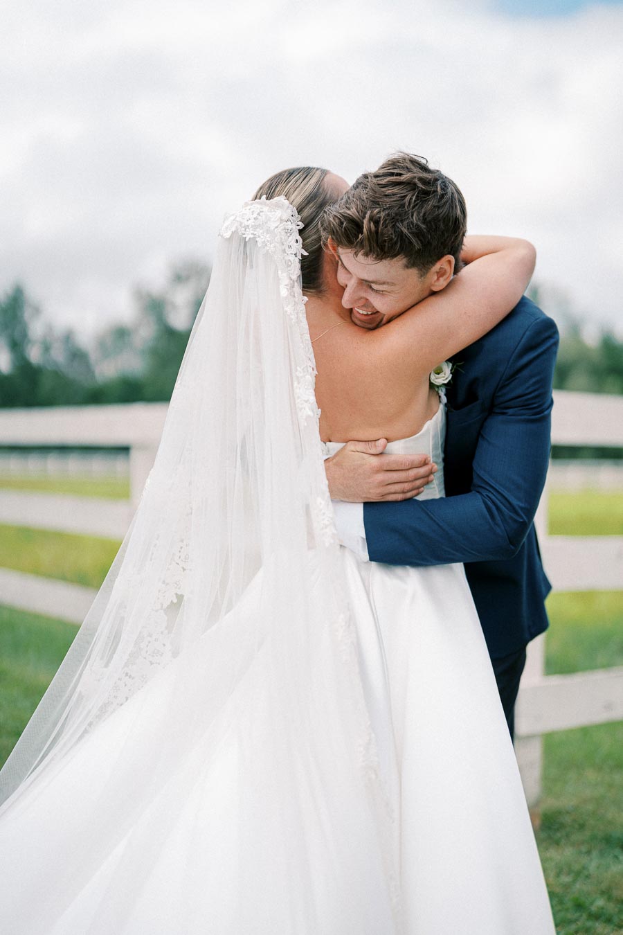 A bride and groom embracing on their wedding day, with the bride wearing a flowing white dress and veil, and the groom in a dark suit, set against a scenic outdoor background.