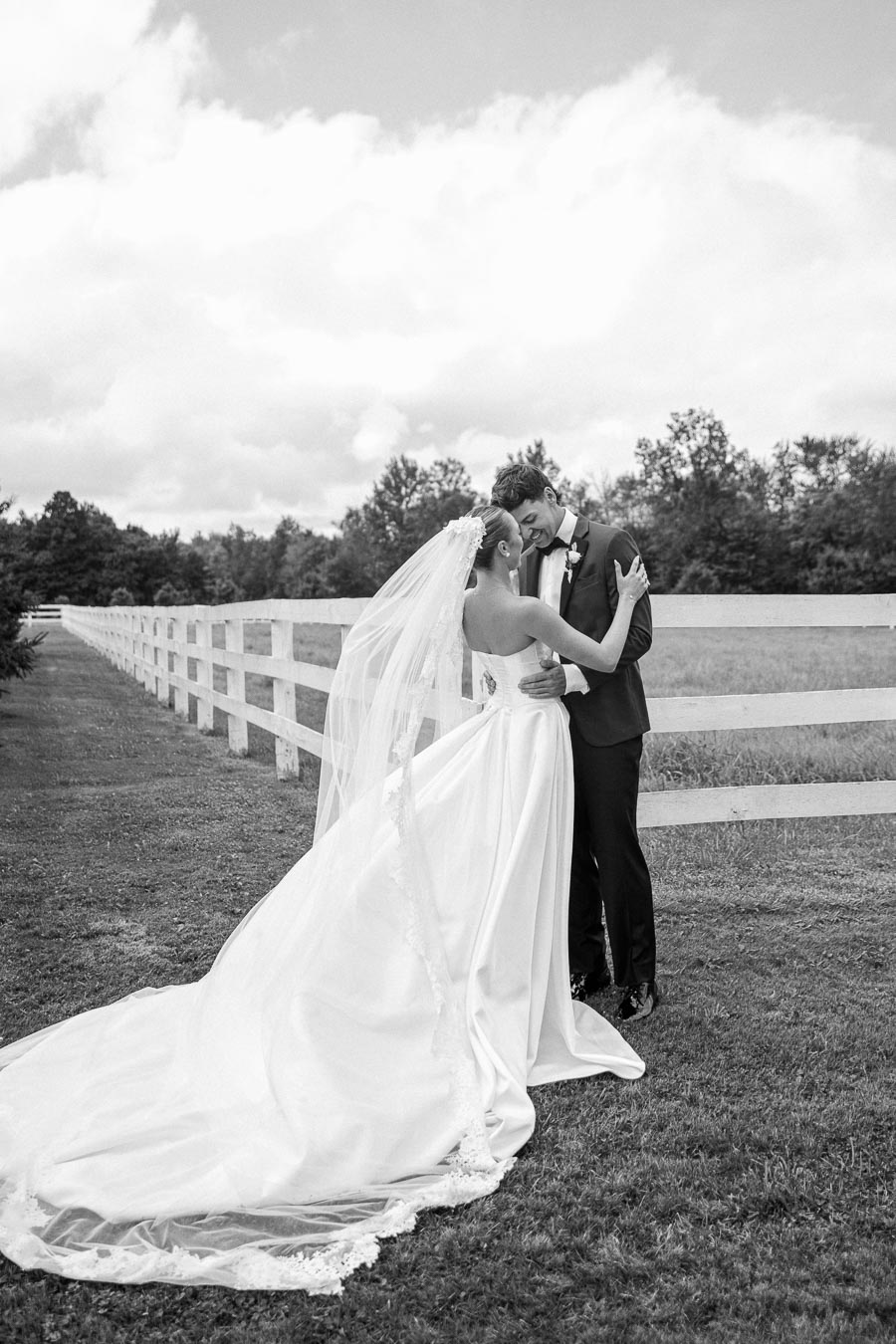 Black and white photo of a bride and groom embracing near a white fence in a picturesque outdoor setting, with the bride in a flowing gown and veil and the groom in a suit, capturing a romantic wedding moment.