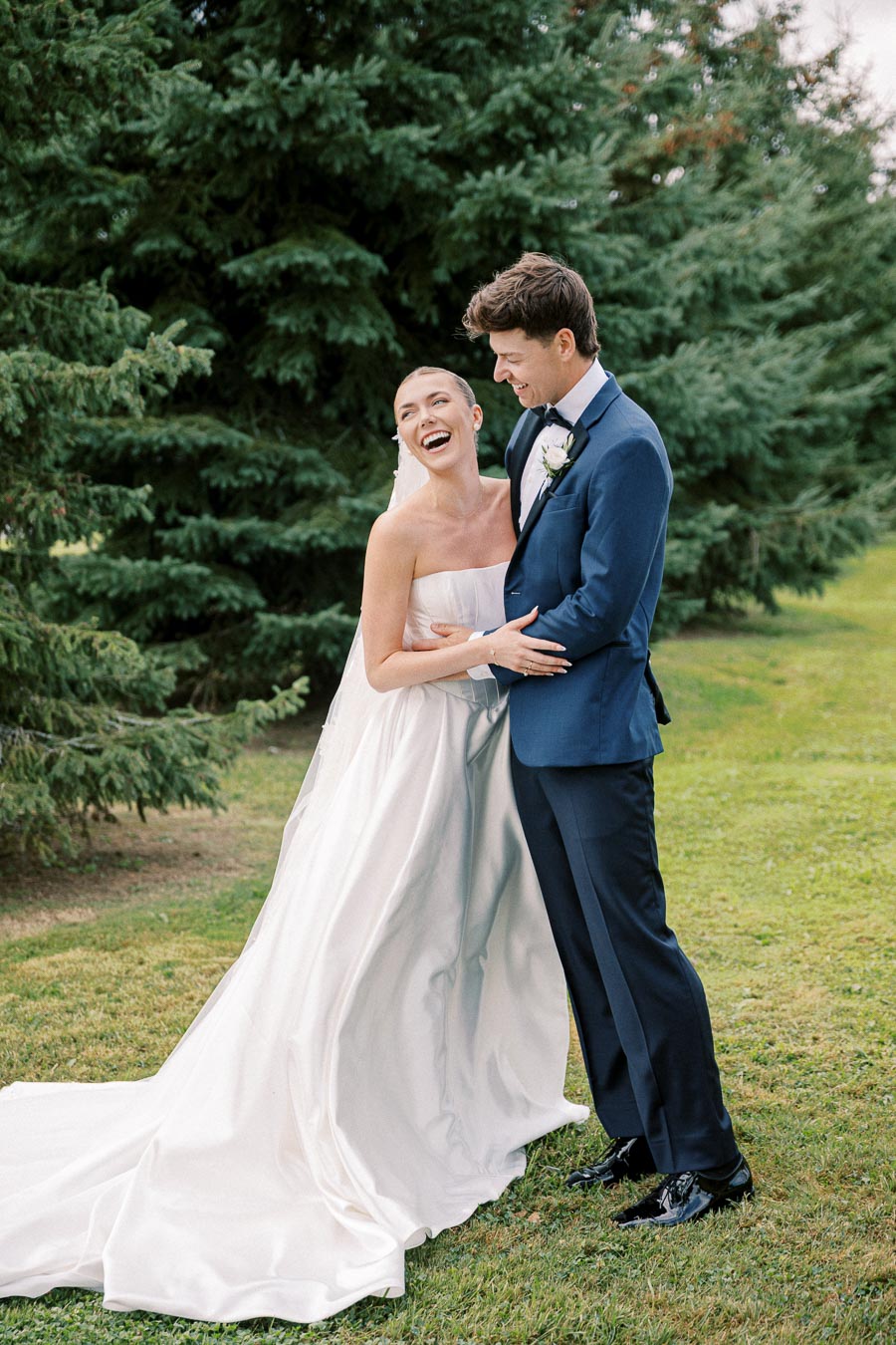 Bride and groom laughing joyfully outdoors in a garden setting, surrounded by lush green trees, with the bride wearing a flowing white gown and the groom in a navy suit and bow tie.