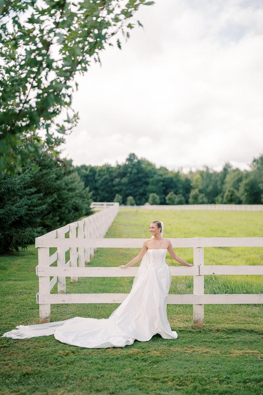 Bride in an elegant white wedding dress stands by a rustic wooden fence in a picturesque green pasture, surrounded by lush trees, under a bright and cloudy sky.