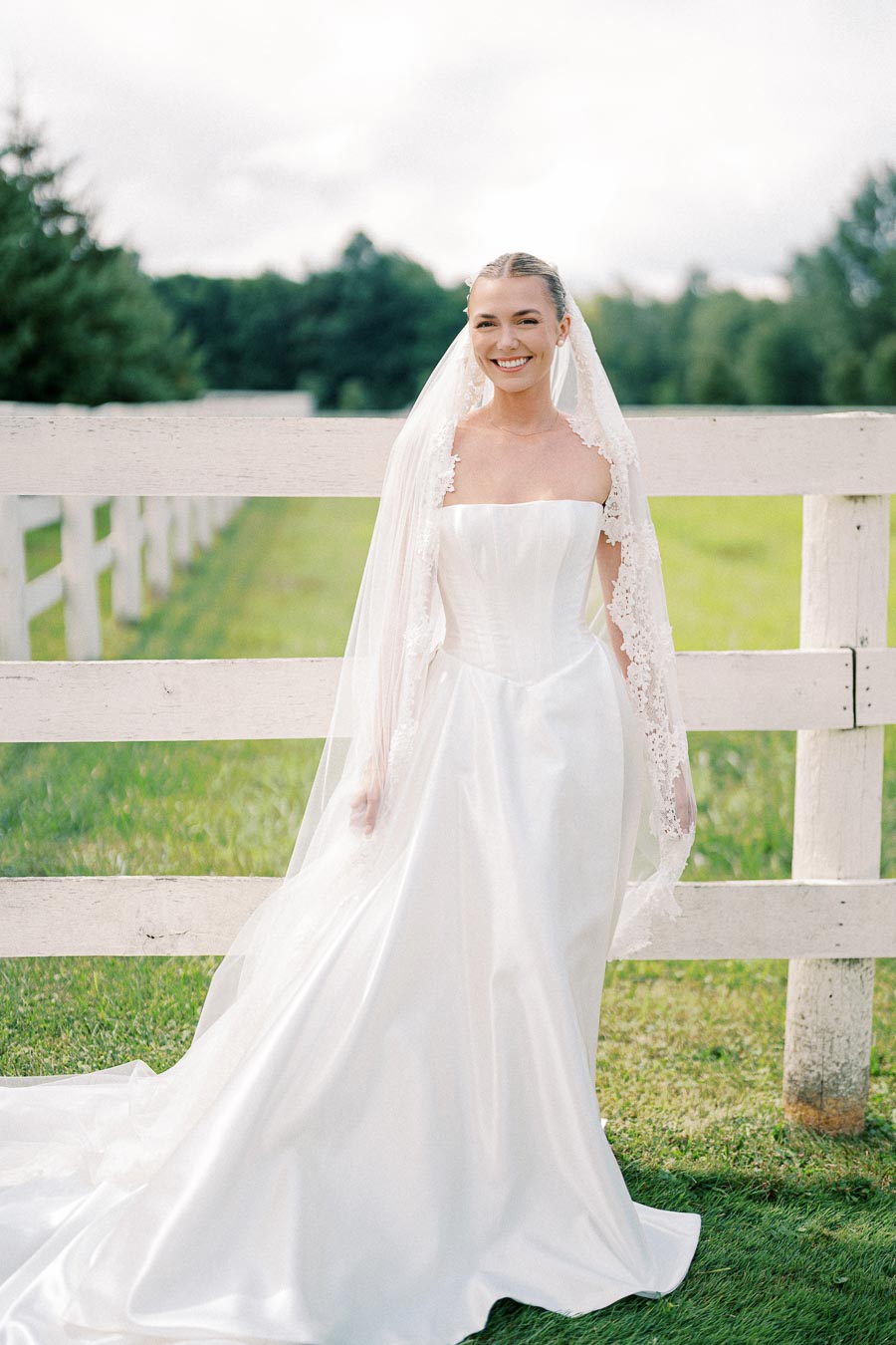 A bride in a white wedding dress with a lace veil stands smiling by a wooden fence, surrounded by lush green grass and trees in a sunlit outdoor setting.