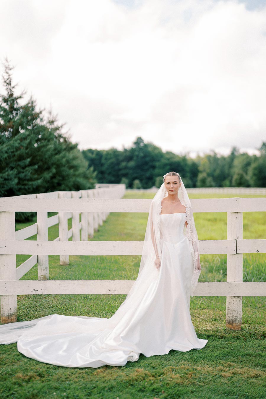 Bride in elegant white wedding gown standing by a rustic white fence in a picturesque countryside setting, with lush green grass and trees in the background.