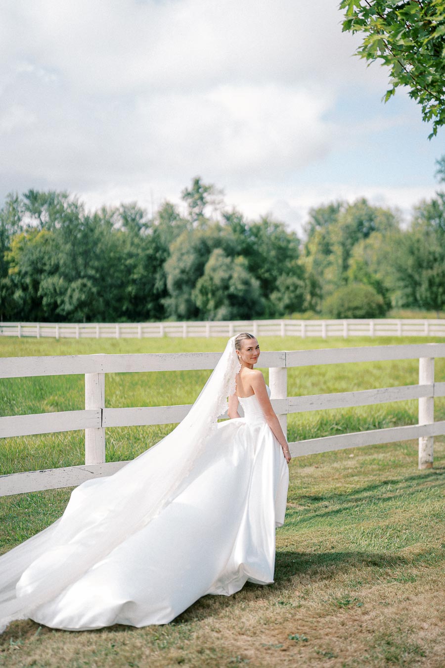 Bride in elegant white wedding dress with a long train and veil stands near a rustic white wooden fence in a sunny green field, surrounded by trees and a partly cloudy sky.