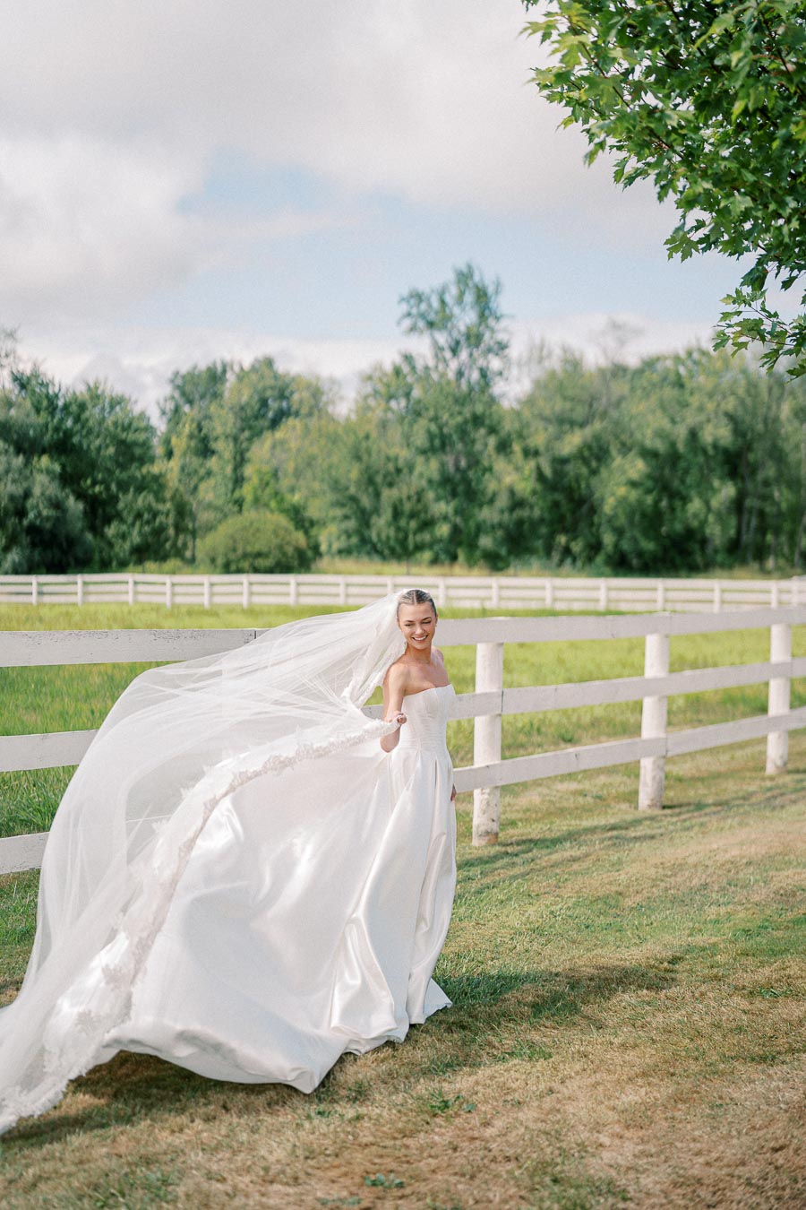 Bride in an elegant white wedding gown with a flowing veil posing outdoors in a lush green field by a wooden fence.