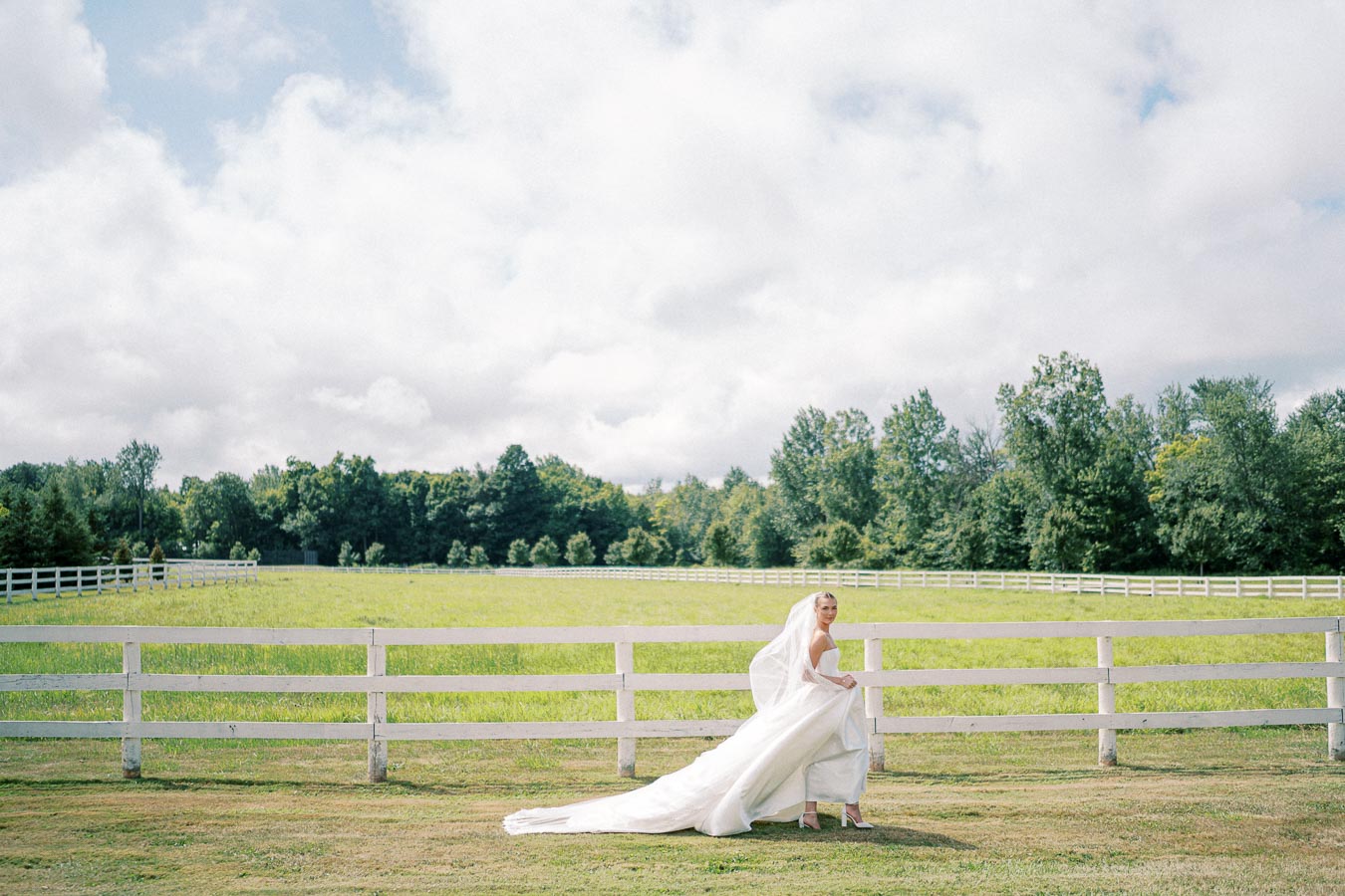 A bride in a white wedding dress with a long train stands by a wooden fence in a scenic green field with trees and a partly cloudy sky in the background.