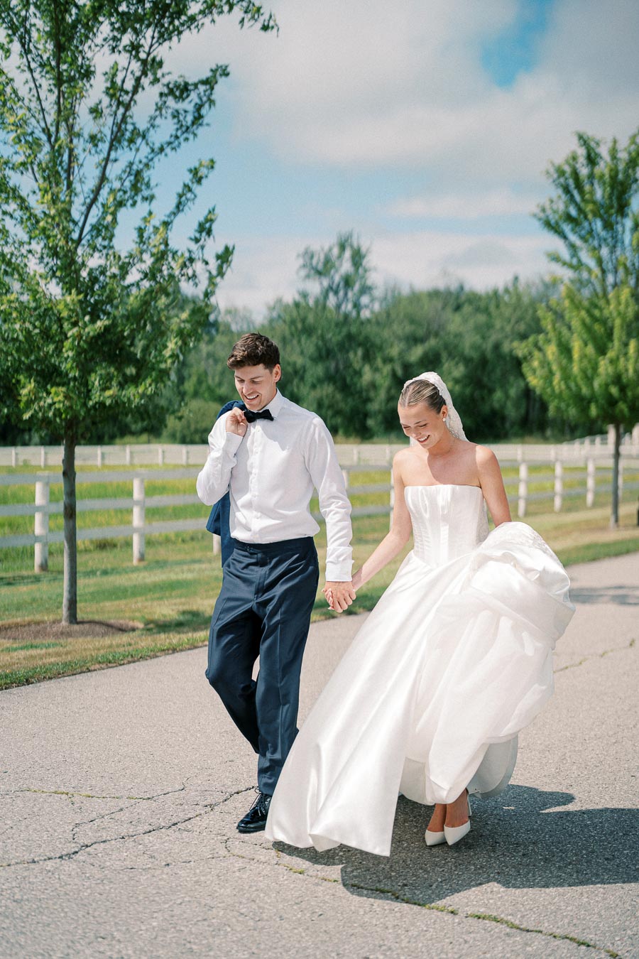A bride and groom walking hand in hand along a tree-lined path, with the bride in a strapless white gown and the groom in a formal attire, capturing a joyous outdoor wedding moment.