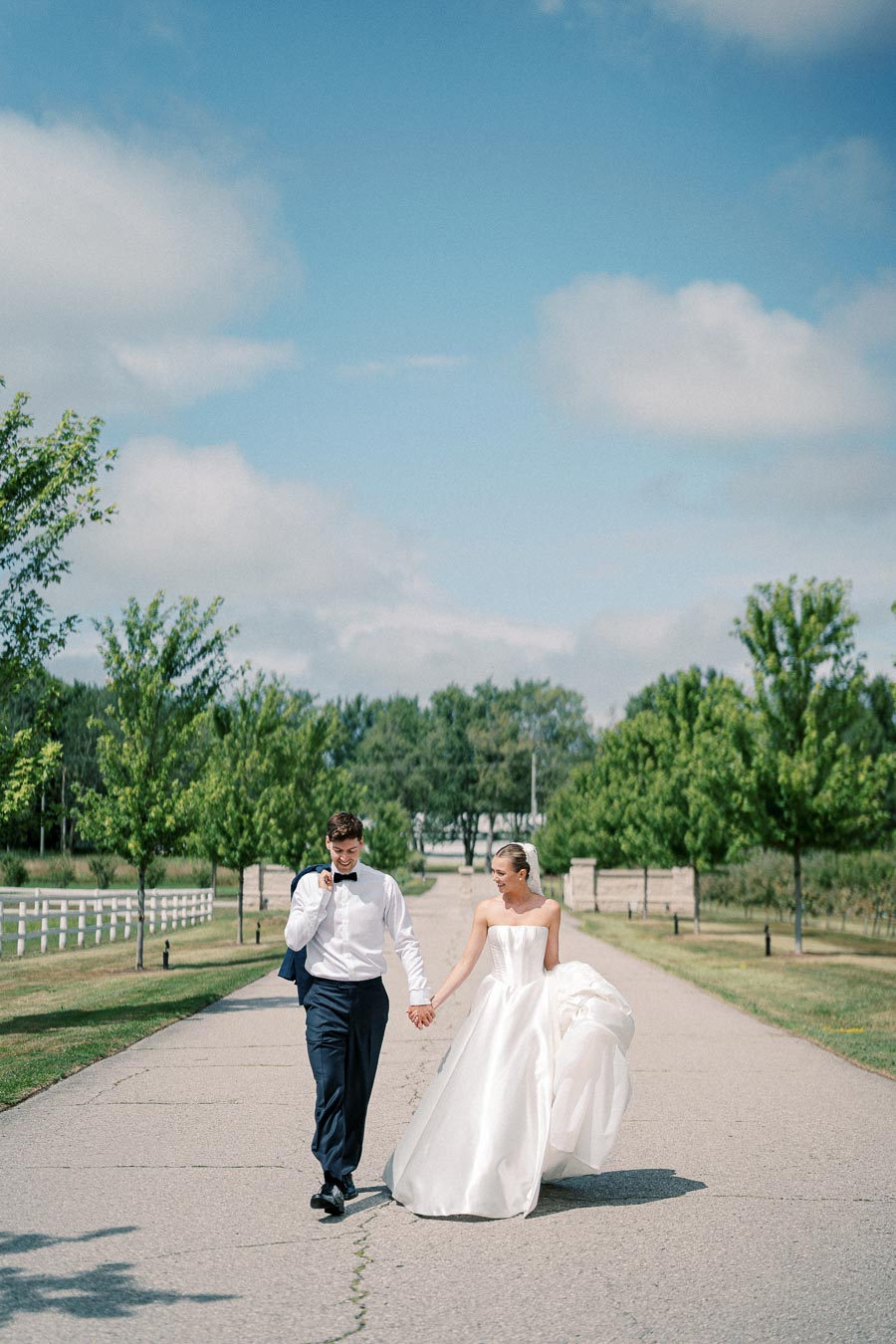 A happy couple walking hand in hand on a tree-lined path during their outdoor wedding, the bride in a flowing white gown and the groom in a tuxedo, with a clear blue sky above.