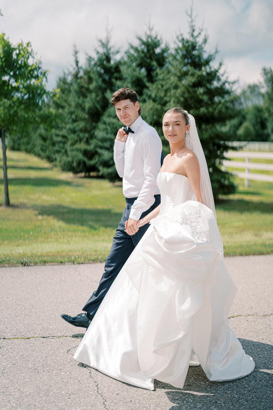 Elegant bride and groom walking outdoors, holding hands on a sunny day with lush green trees in the background, bride in a strapless white gown and groom in a classic white shirt and dark trousers.