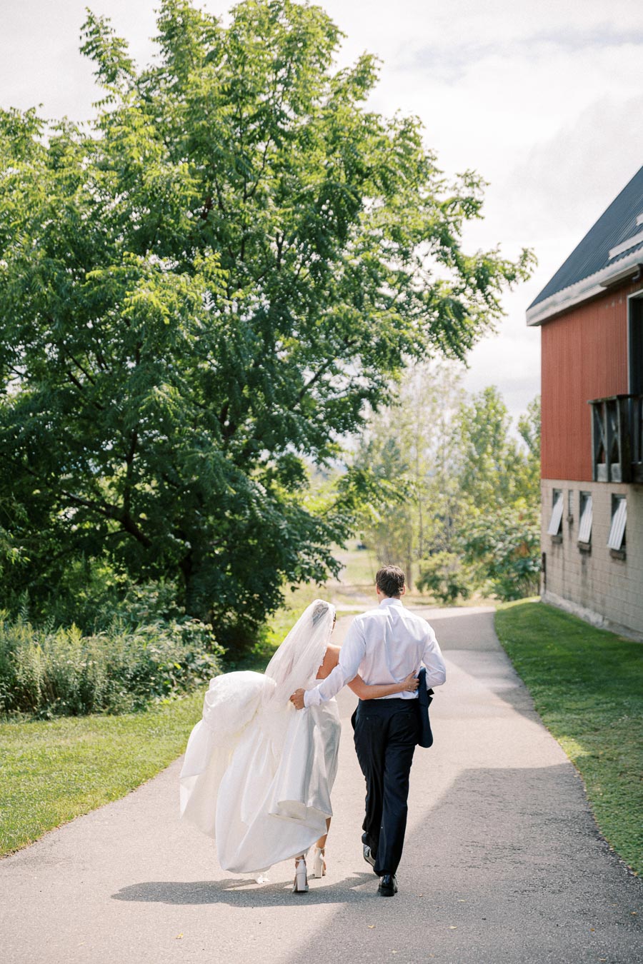 Bride and groom walking hand in hand down a tree-lined path on a sunny wedding day, with a rustic barn in the background.