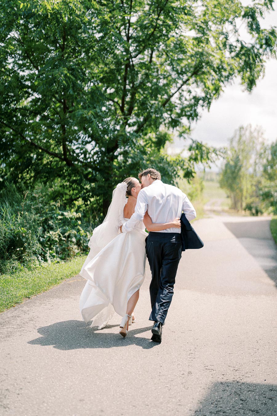 A bride and groom joyfully walking hand in hand on a sunlit path with lush greenery in the background.