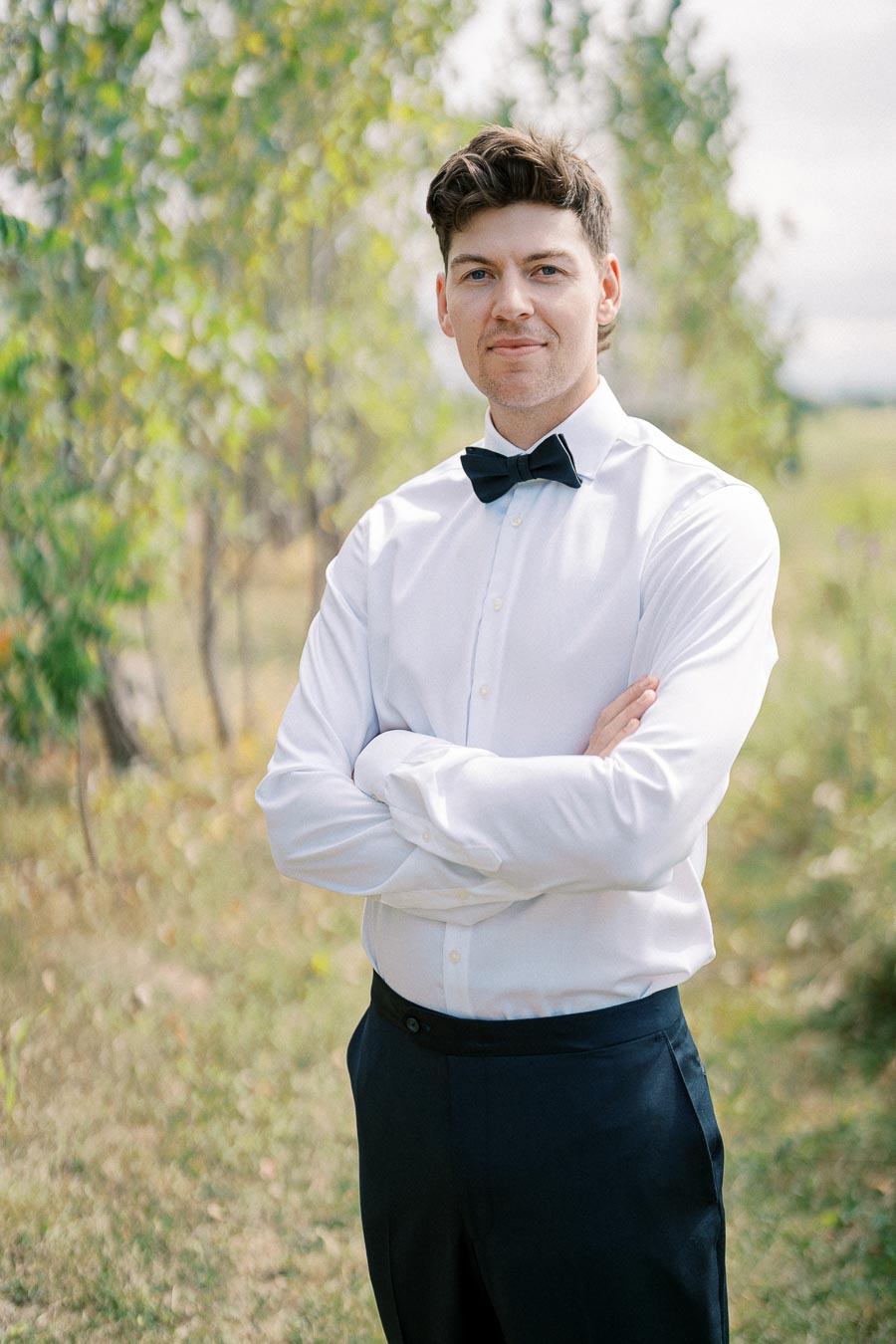 A man wearing a white dress shirt and black bow tie standing outdoors with arms crossed, smiling confidently in a sunlit field with green trees in the background.
