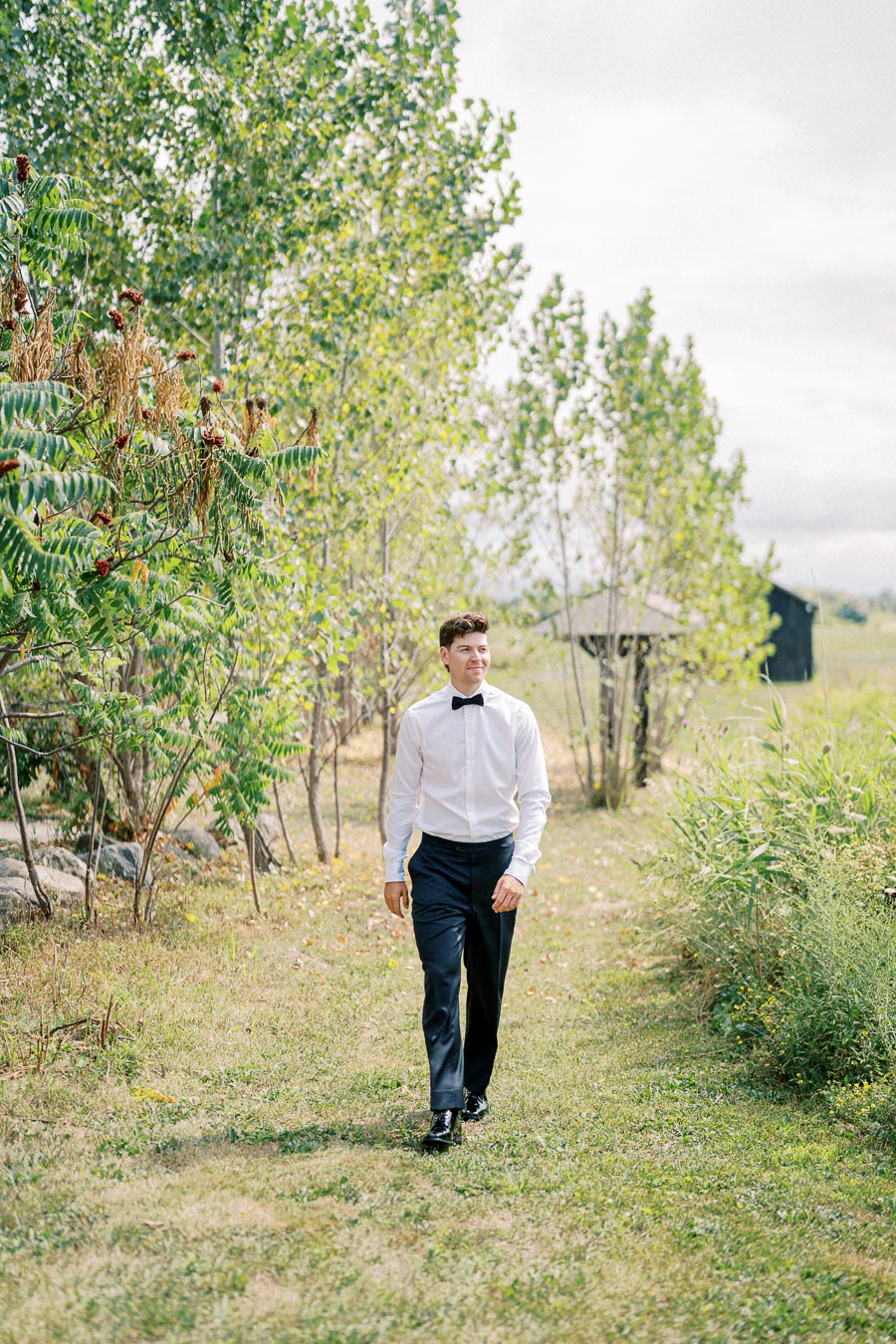 A man in a formal outfit, wearing a white shirt and black bow tie, walks along a grassy path lined with green trees and shrubs, under a cloudy sky.