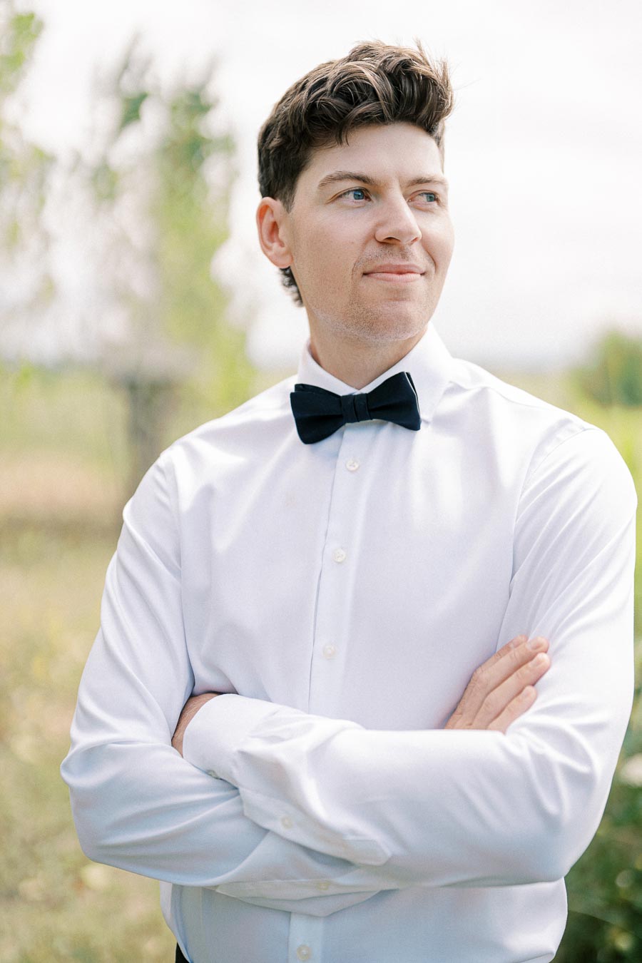 A man wearing a white shirt and black bow tie stands outdoors with arms crossed, looking to the side.