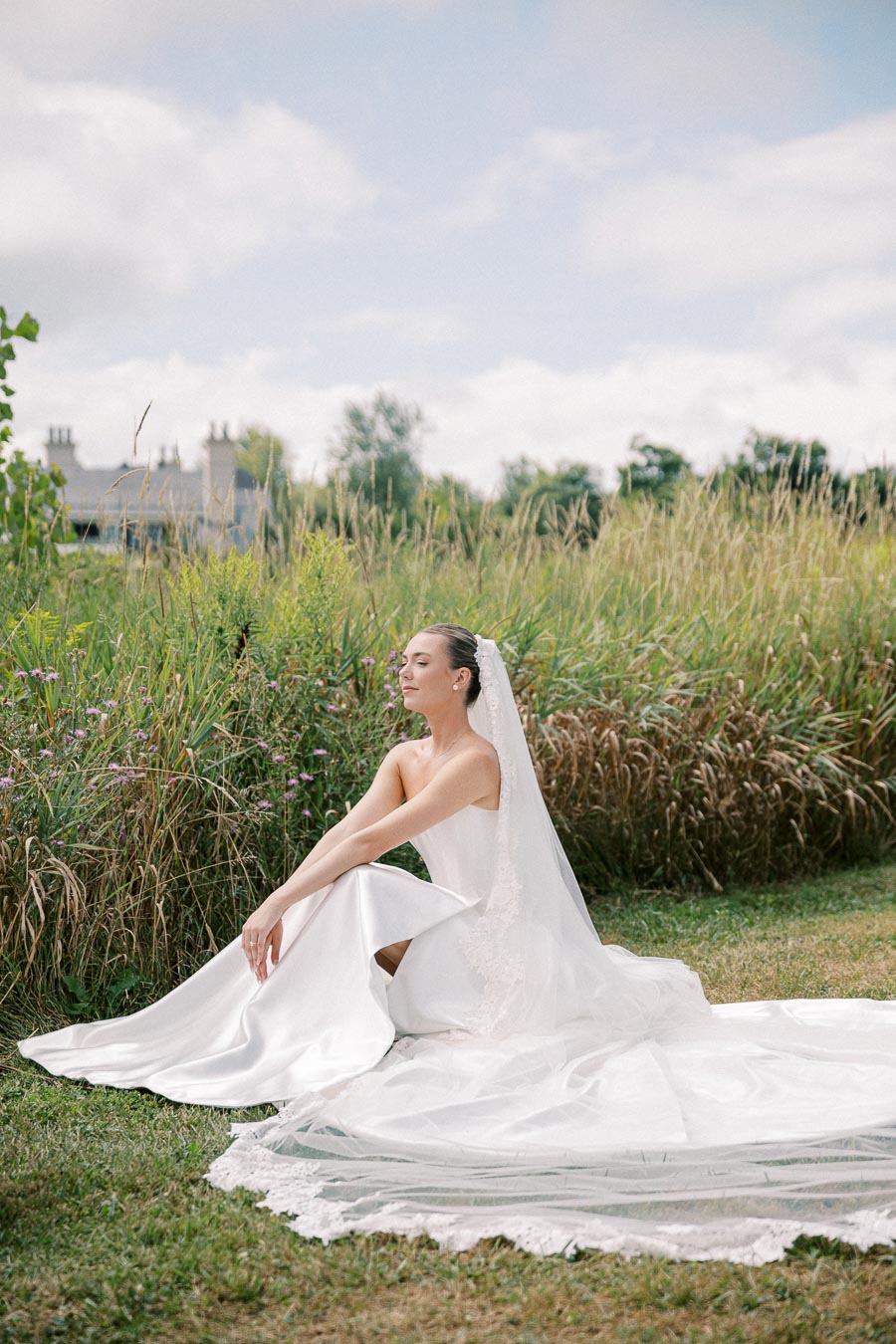 Bride in elegant white wedding dress and veil sitting gracefully on a grassy field, surrounded by lush greenery and wildflowers under a partly cloudy sky.