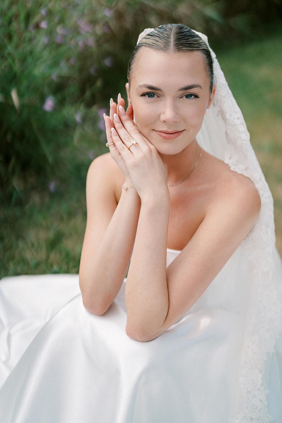 A bride in a white wedding dress and veil smiles softly, with hands gently touching her face, showcasing an elegant ring, against a blurred natural background.