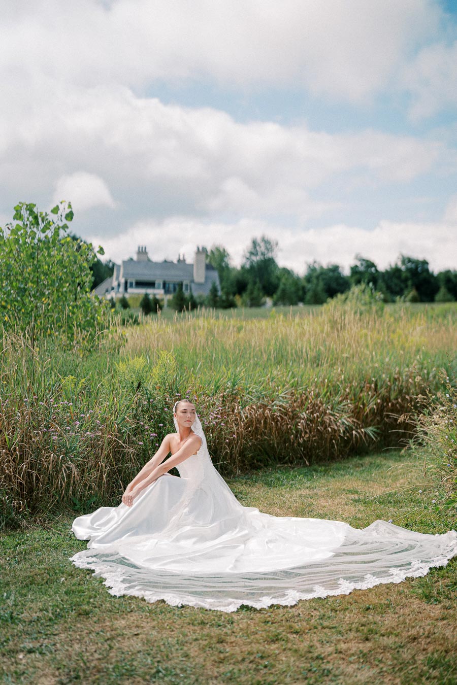 Bride in elegant white wedding dress with long train sitting in a picturesque meadow, with a charming country house in the background under a cloudy sky.