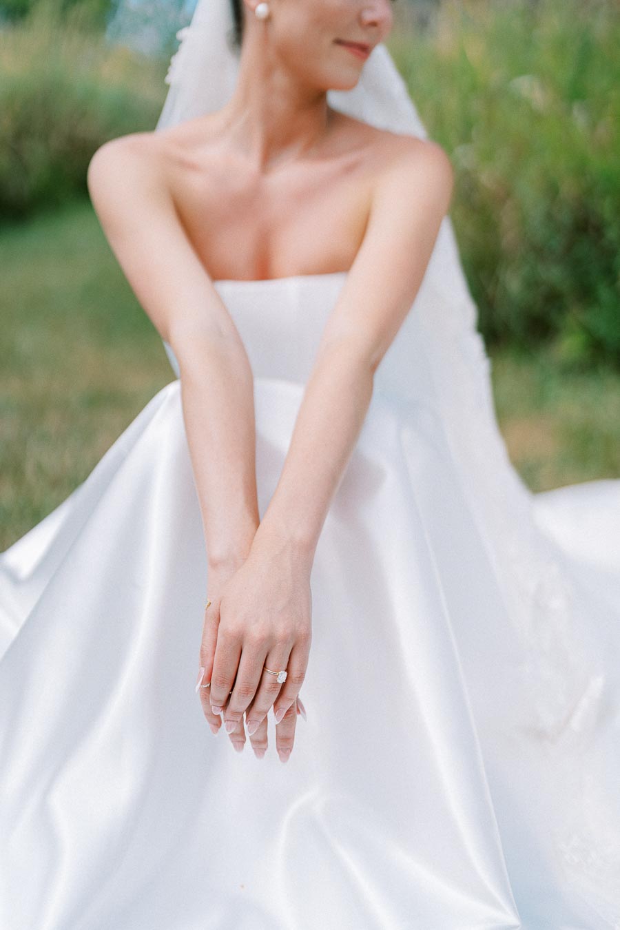 A bride in a white strapless wedding gown sitting on grass, showing her hands with an elegant diamond engagement ring.