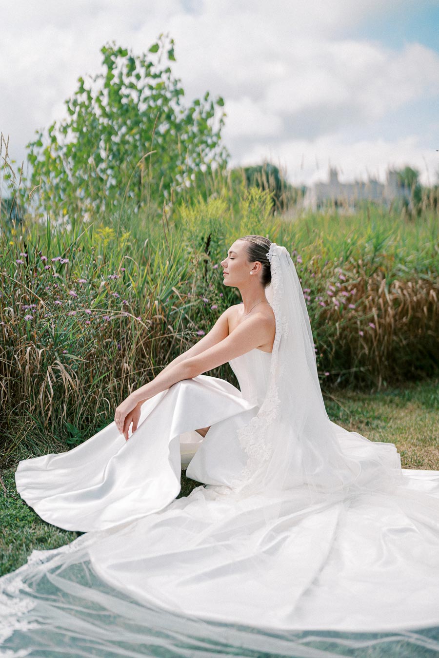 A bride in a flowing white wedding gown and veil sits gracefully on the grass against a backdrop of lush greenery and cloudy sky.