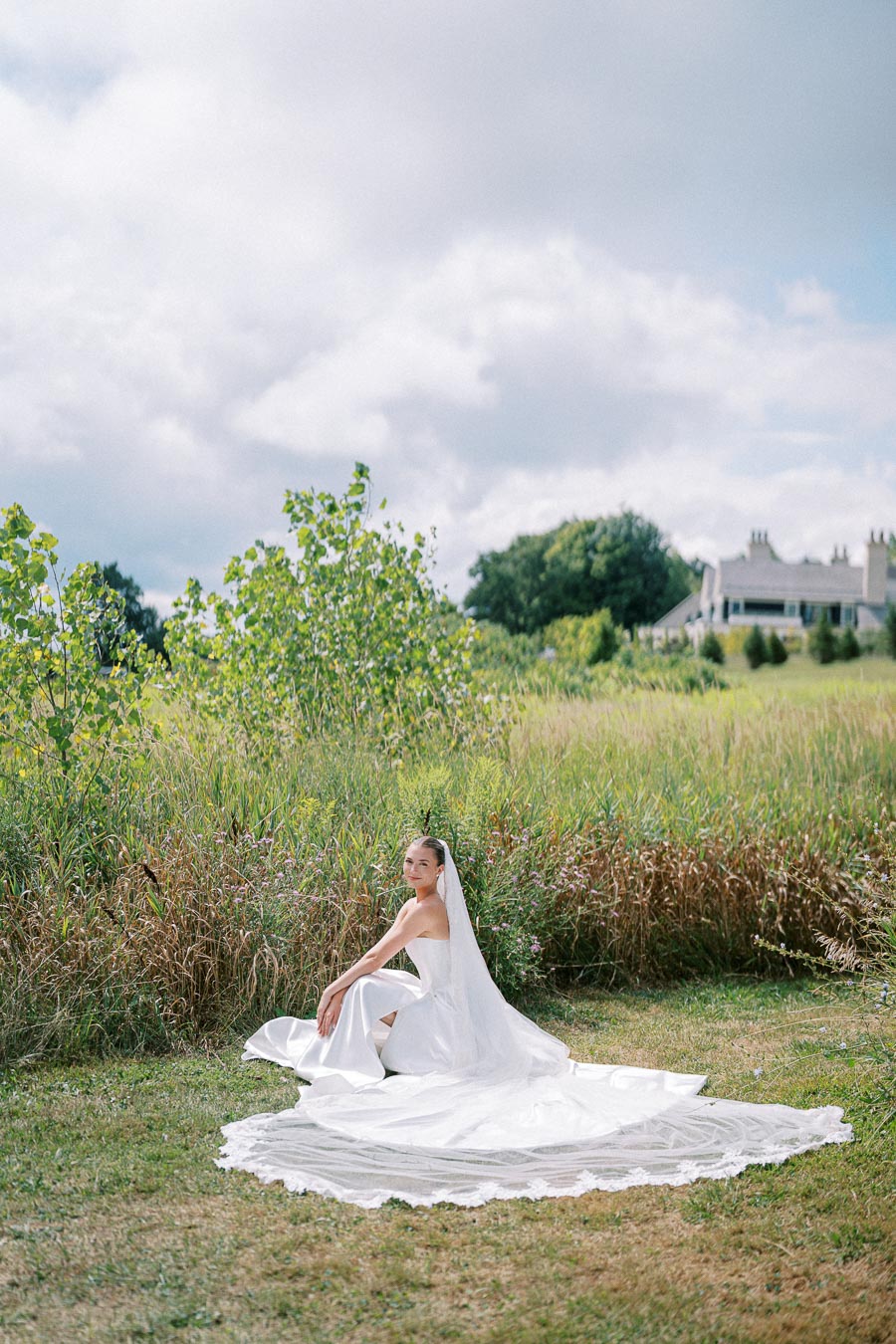 Bride in elegant white wedding dress and veil sitting on grassy field with a scenic countryside backdrop.