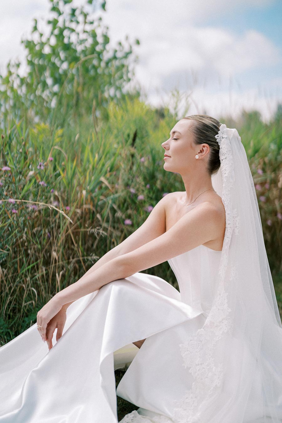 Elegant bride in a white wedding dress and veil sitting peacefully in a lush green garden, exuding serenity and grace under a partly cloudy sky.