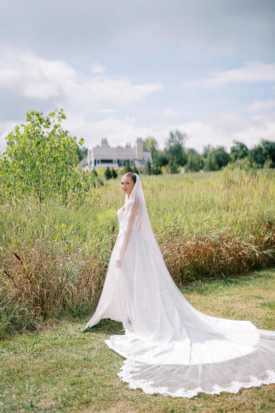 A bride in an elegant white wedding dress and veil stands in a grassy field, with a large country house in the background under a partly cloudy sky.