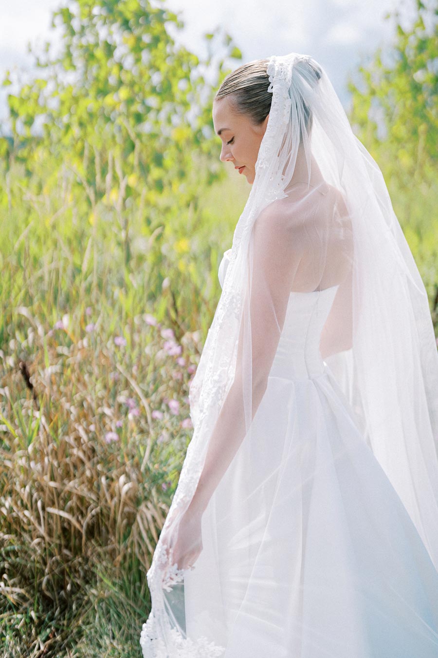 A bride in a white wedding dress and veil stands outdoors, surrounded by lush greenery and wildflowers, embracing a serene moment in nature.
