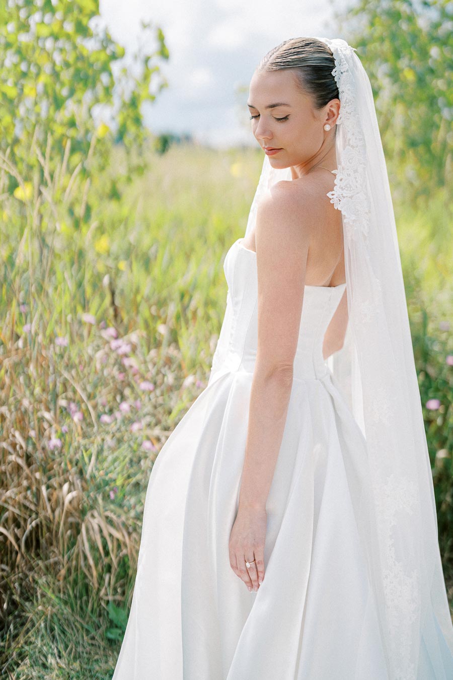 Bride in elegant white wedding dress and lace veil posed in a serene garden setting.