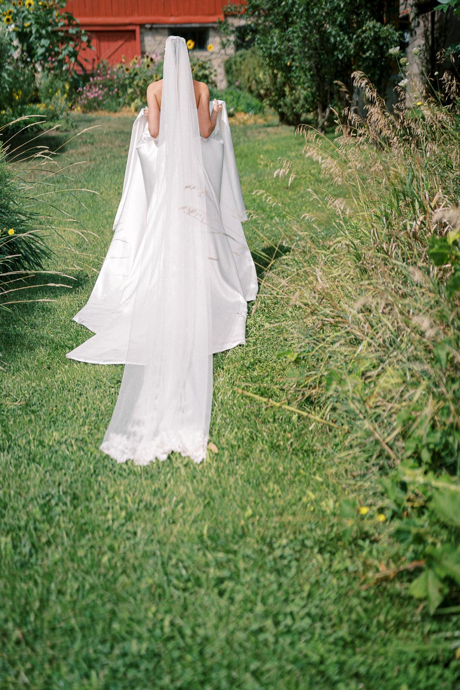 Bride walking on a grassy path, wearing an elegant white wedding dress with a long veil, surrounded by lush greenery and flowers.