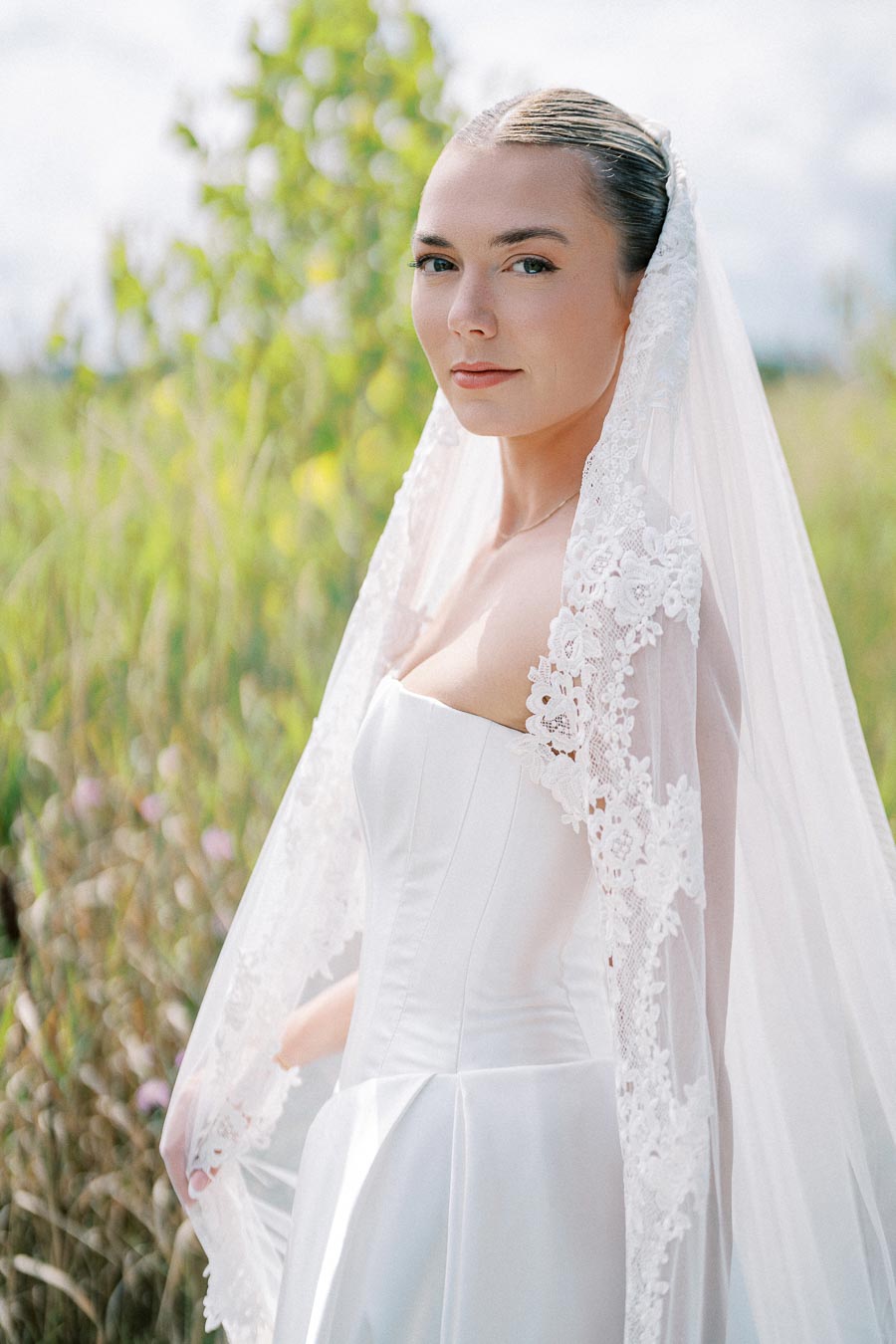 Elegant bride in a white wedding dress with a lace veil, standing outdoors with a serene expression, surrounded by lush greenery.