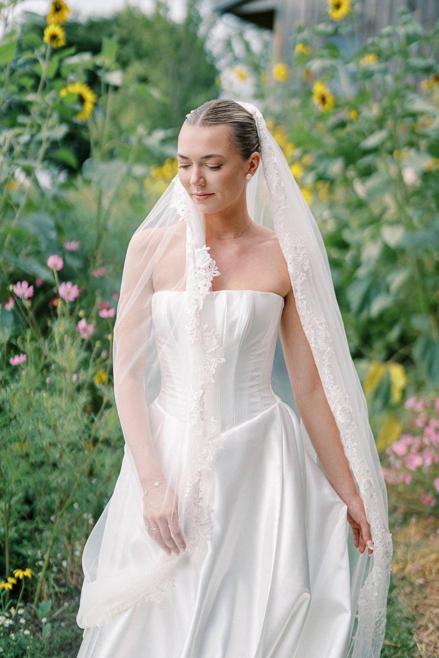 Elegant bride in a strapless white wedding gown with lace veil, standing in a lush garden with sunflowers and pink flowers.
