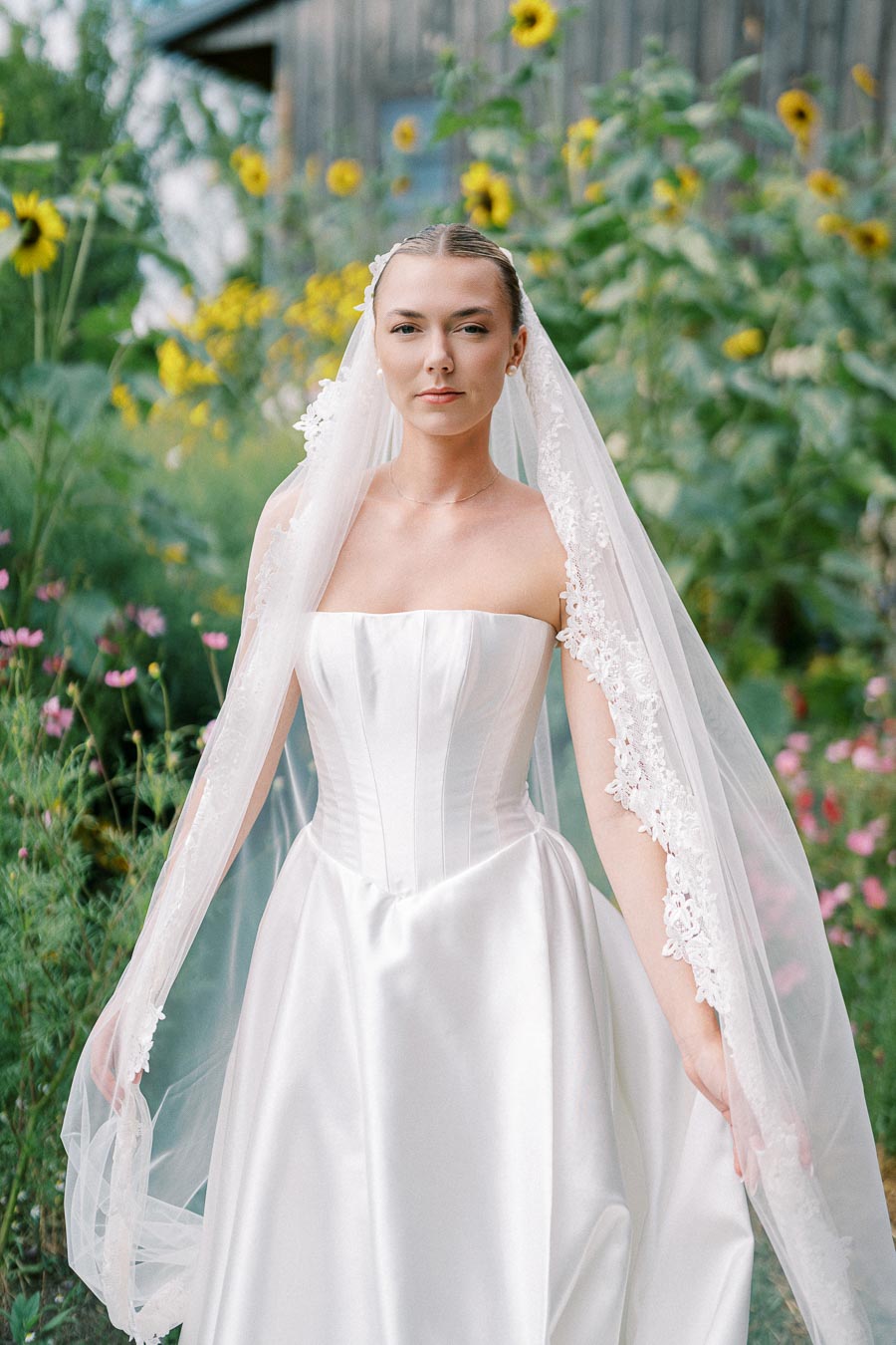 Bride in a strapless satin wedding gown with lace veil stands in a garden surrounded by sunflowers and greenery.