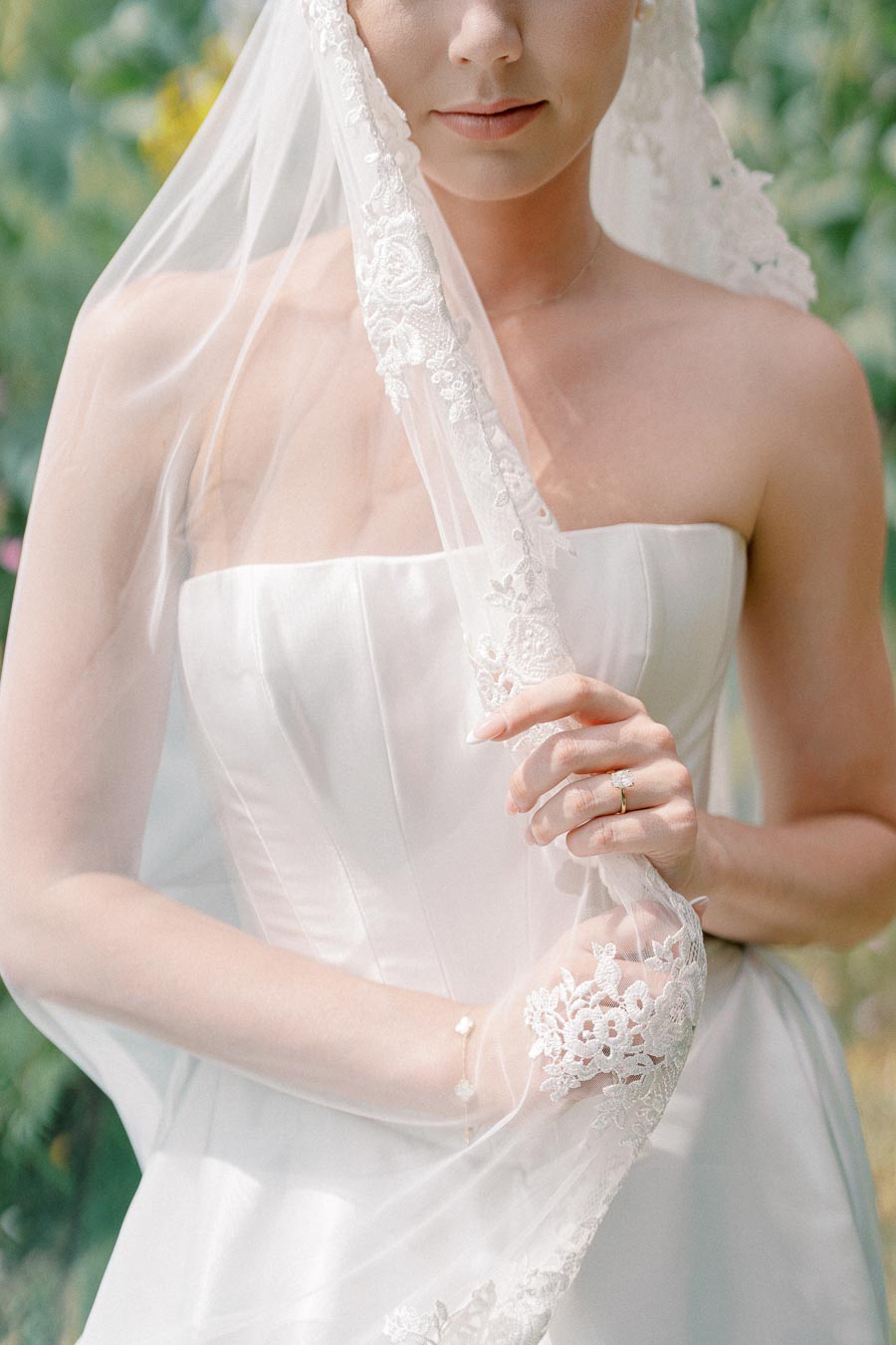 A bride in a strapless white wedding dress holds her lace veil, showcasing a diamond ring, in a serene outdoor setting.