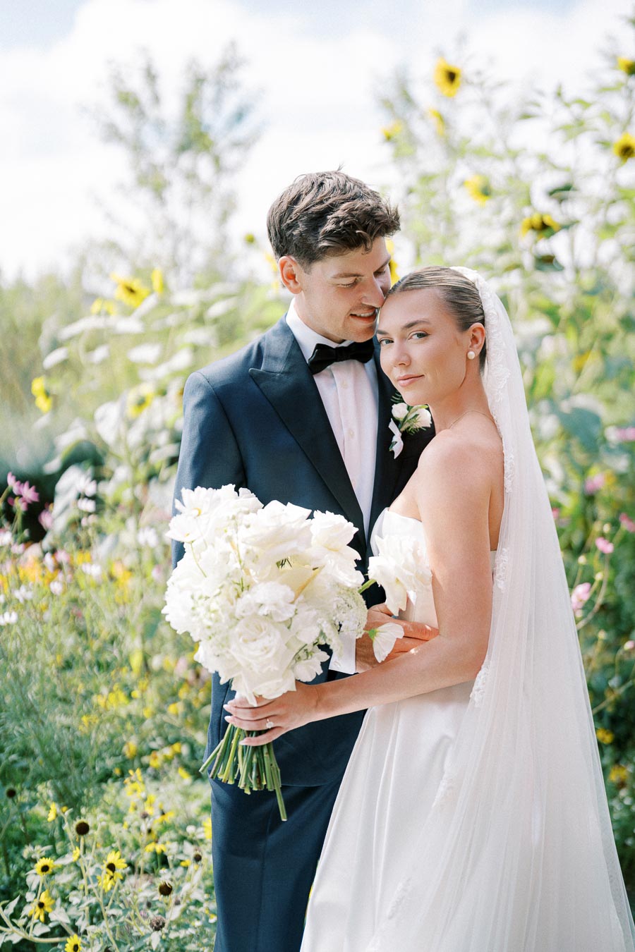 A bride and groom embracing in a sunlit garden setting, with the bride holding a bouquet of white flowers and sunflowers in the background.