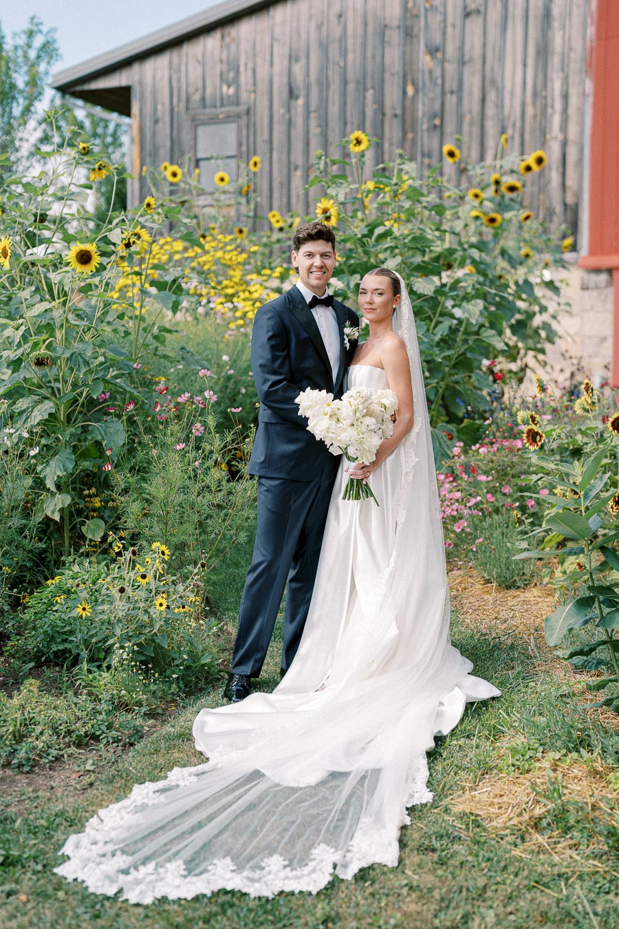 Elegant bride and groom standing in a vibrant garden, surrounded by sunflowers and wildflowers, with a rustic wooden barn in the background.